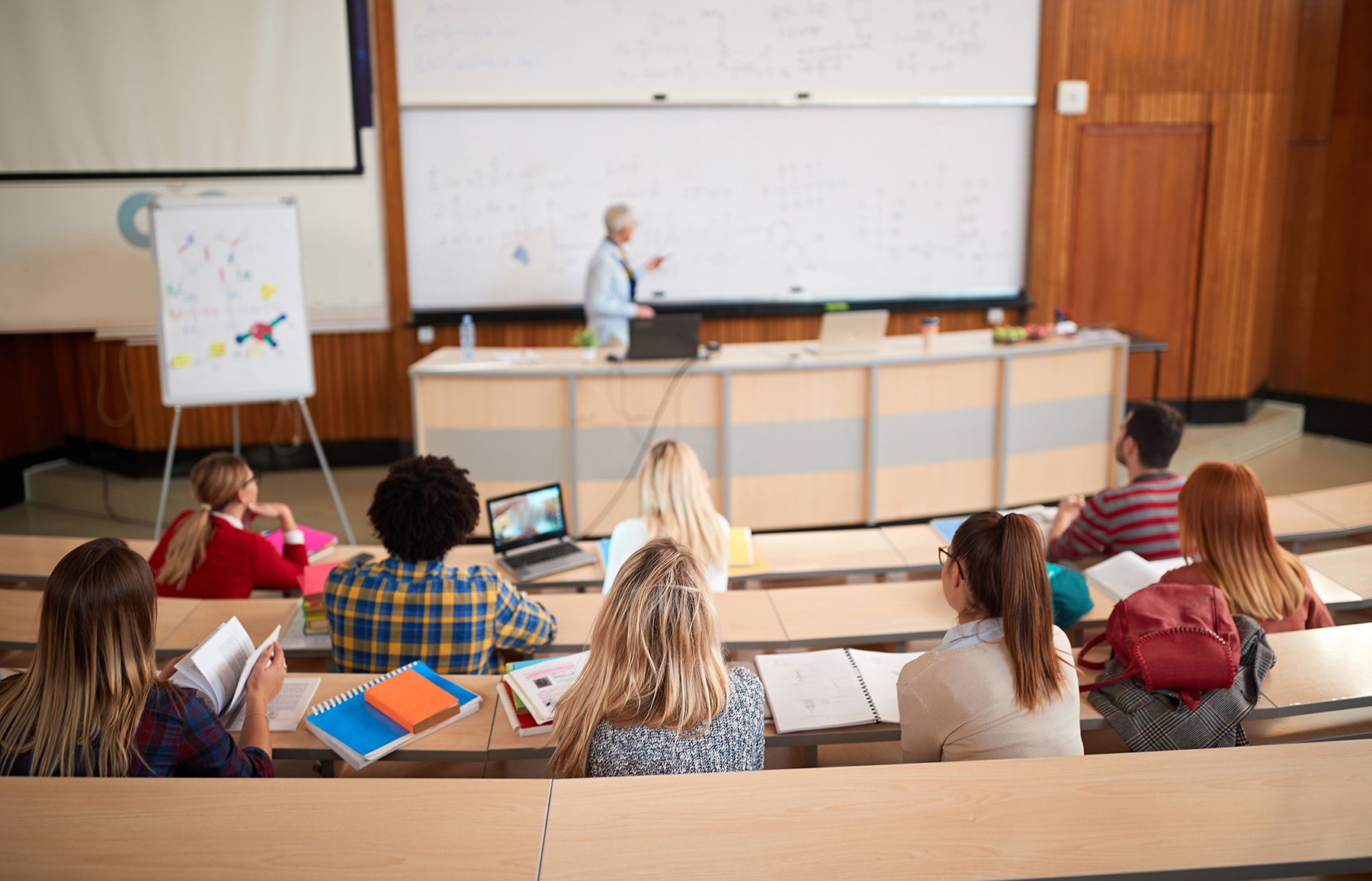 Students attending a lecture in a university classroom, listening to a professor at the whiteboard, representing academic instruction.